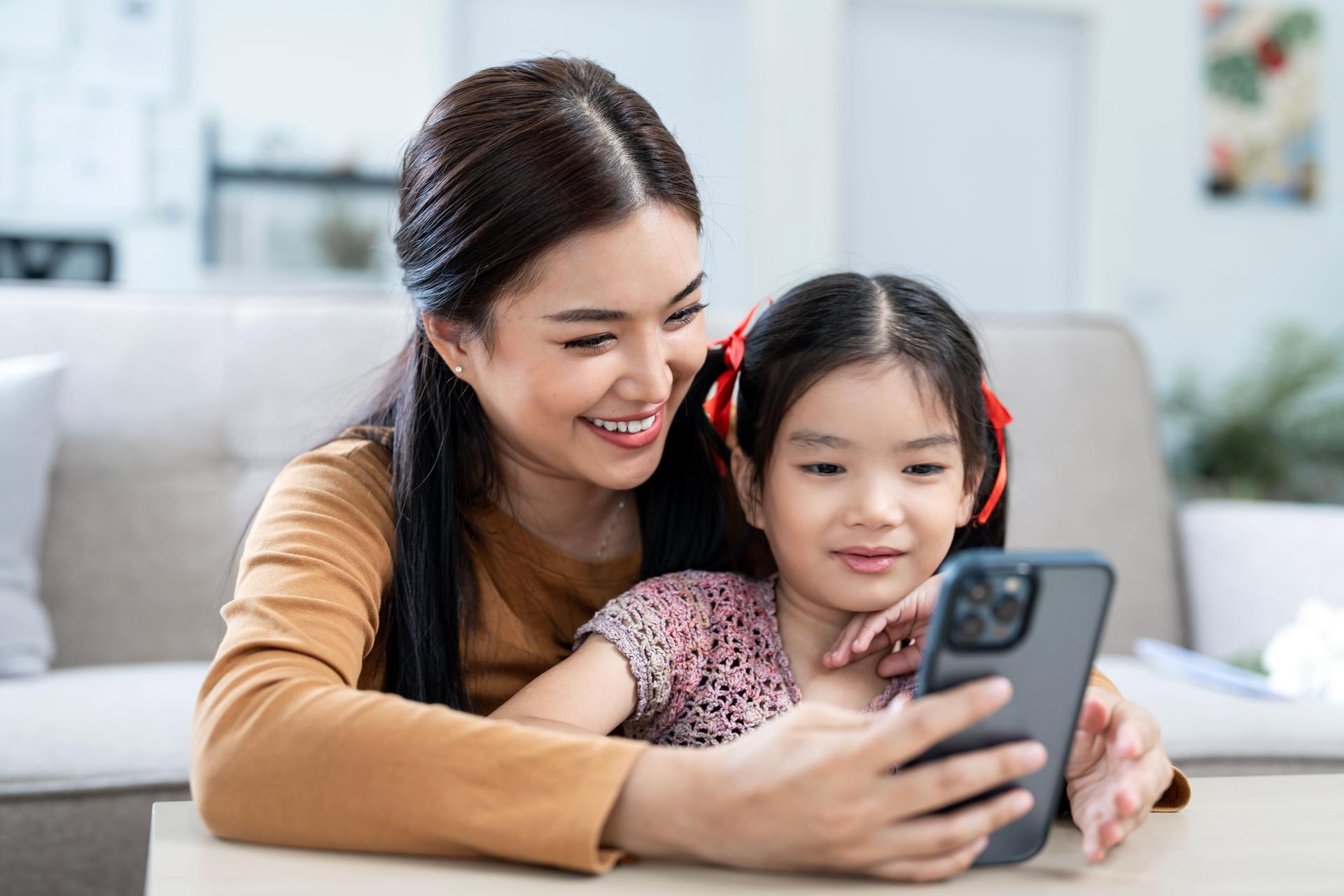Happy family, beautiful mom and her daughter spending happy time together at home, using mobile phone for elearning, playing games or streaming video on movie website.